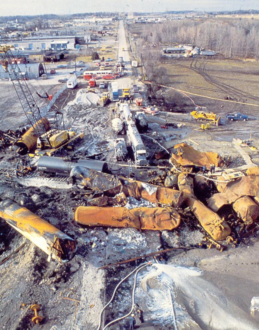 Colour aerial photograph of the Mississauga derailment aftermath showing twisted orange and yellow tanker cars scattered across Mavis Road, with cleanup vehicles and emergency equipment