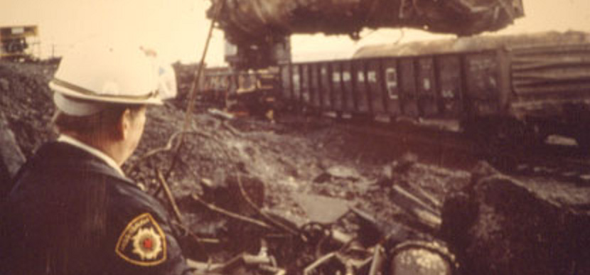Police officer surveying the Mississauga derailment cleanup, with a twisted tanker car overhead and debris scattered across the site